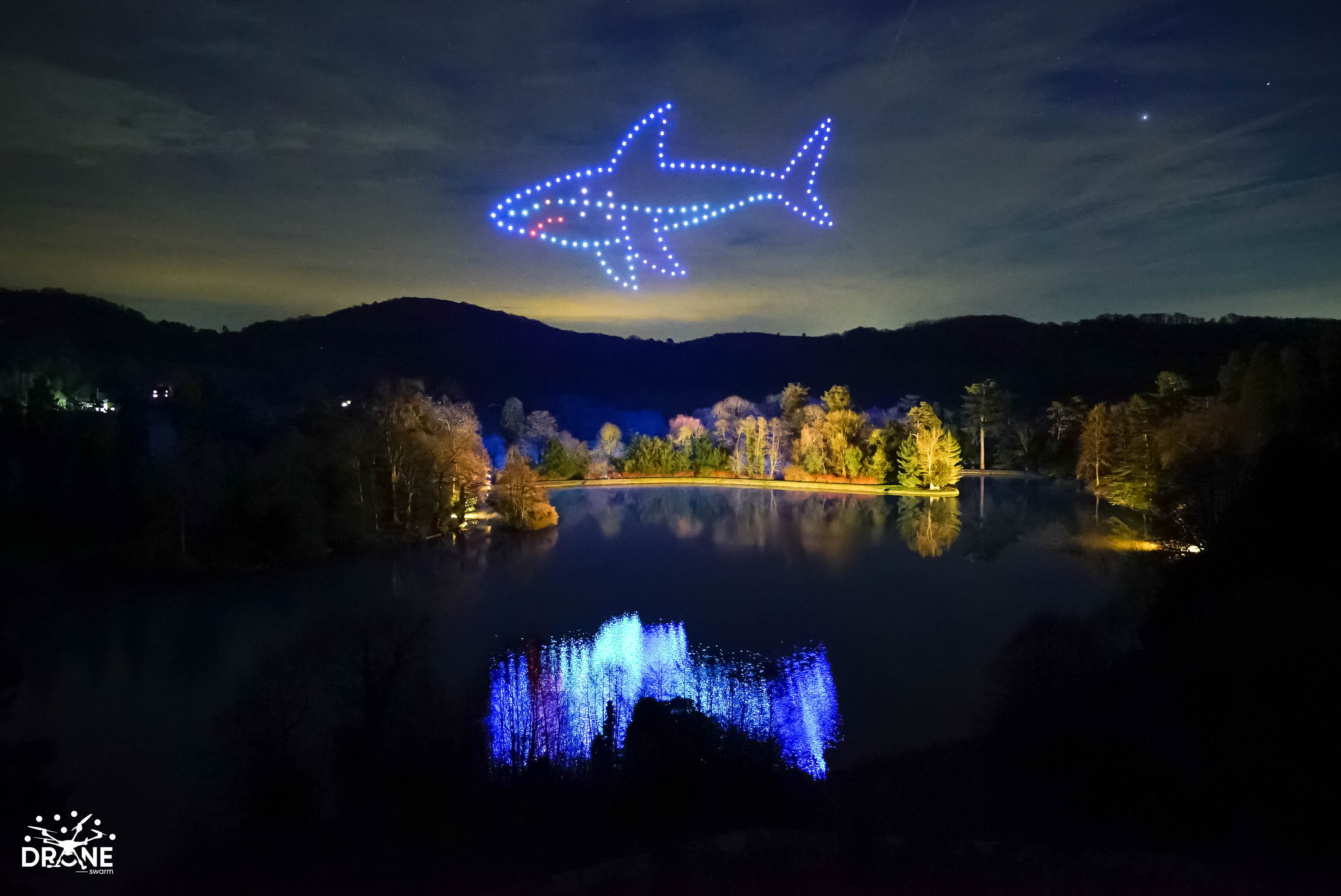 A shark-shaped drone formation over a lake, with the lights reflected in the water